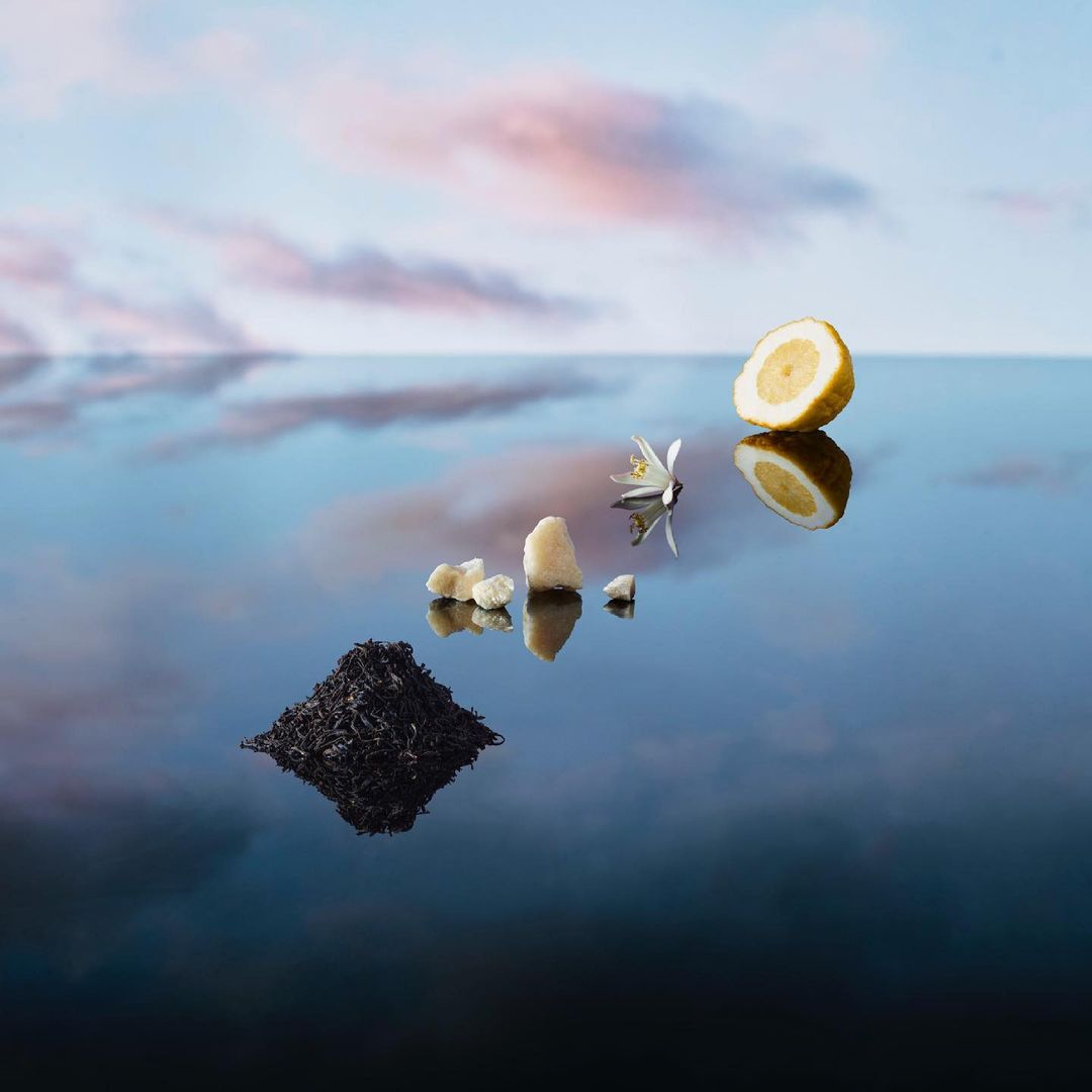 Lemon slice, sugar, and tea leaves are arranged on a reflective surface against a serene sky backdrop, product photography.
