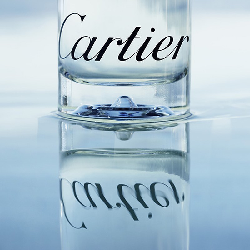 Close-up studio shot of clear glass containing a liquid droplet, featuring the Cartier logo in black against a bright, reflective background.
