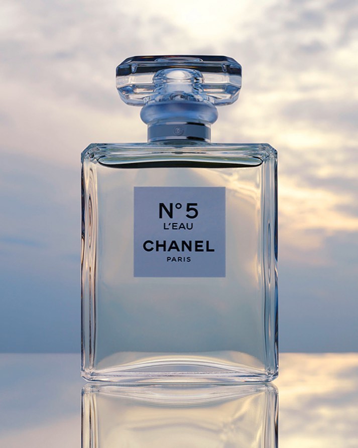 Close-up of a clear Chanel N°5 L'Eau perfume bottle reflecting on a glossy surface with a soft, cloudy sky in the background.