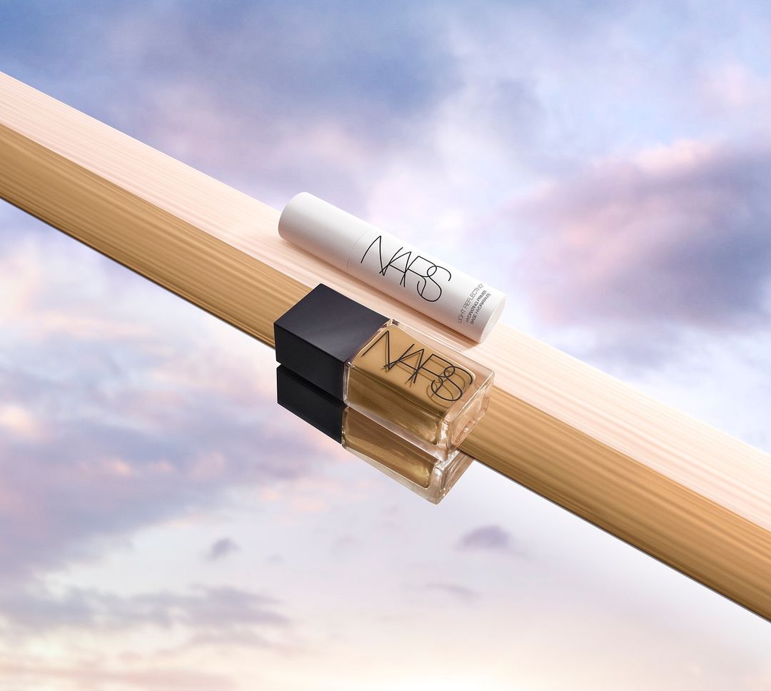 NARS cosmetic foundation and concealer products displayed diagonally on a light wood plank against a pastel cloudy sky background.