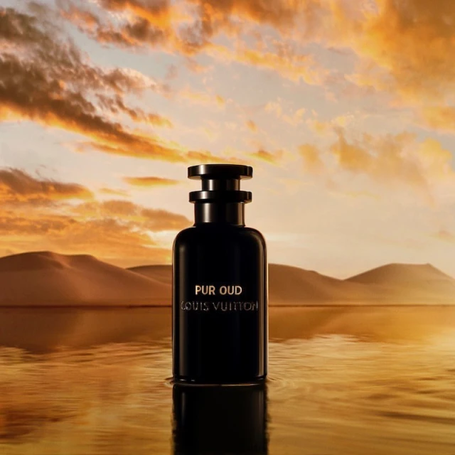 Dark Louis Vuitton perfume bottle on reflective surface with desert hills and orange sunset sky in the background, beauty shot.