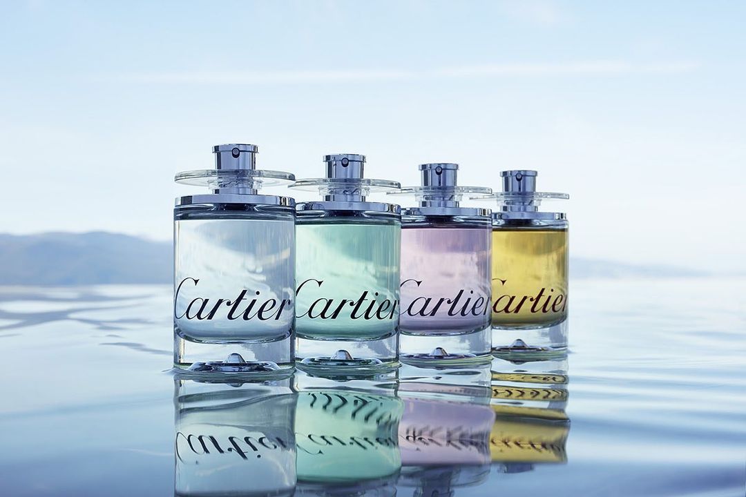 Four clear perfume bottles with silver tops are arranged on a reflective surface, displaying a range of pastel tints against a pale sky backdrop.