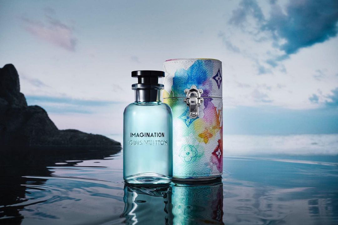 Louis Vuitton Imagination perfume bottle and patterned travel case on reflective surface with blurred shoreline and sky backdrop.