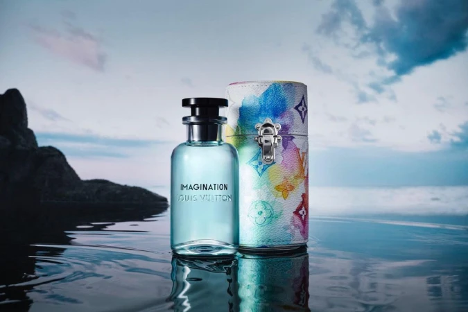 Louis Vuitton Imagination perfume bottle and patterned travel case on reflective surface with blurred shoreline and sky backdrop.