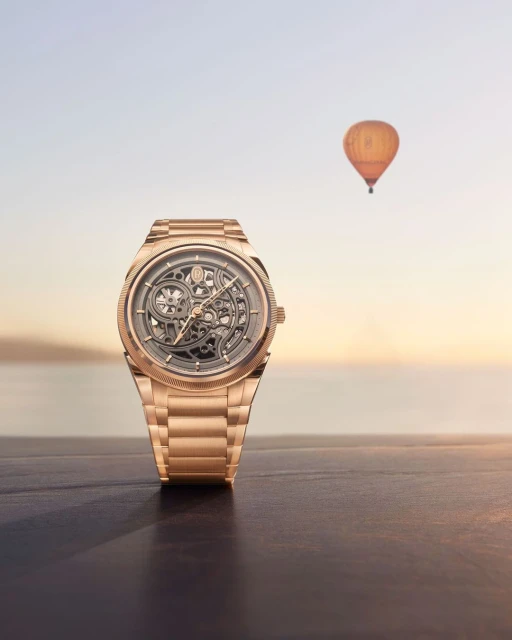 Wristwatch with skeleton dial and gold band on table with hot air balloon in the background at sunset; luxury timekeeping.
