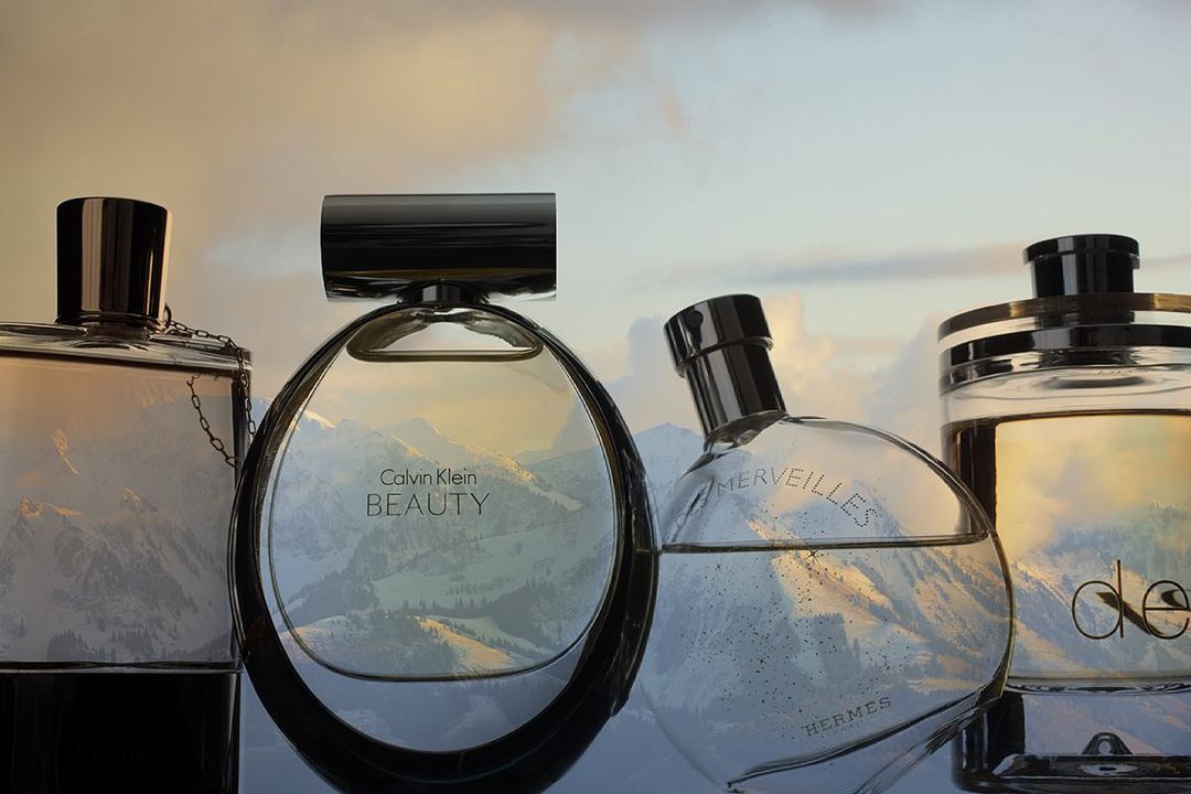 Close-up of glass perfume bottles featuring reflections of a mountain range, set against a soft, dreamy sky backdrop.