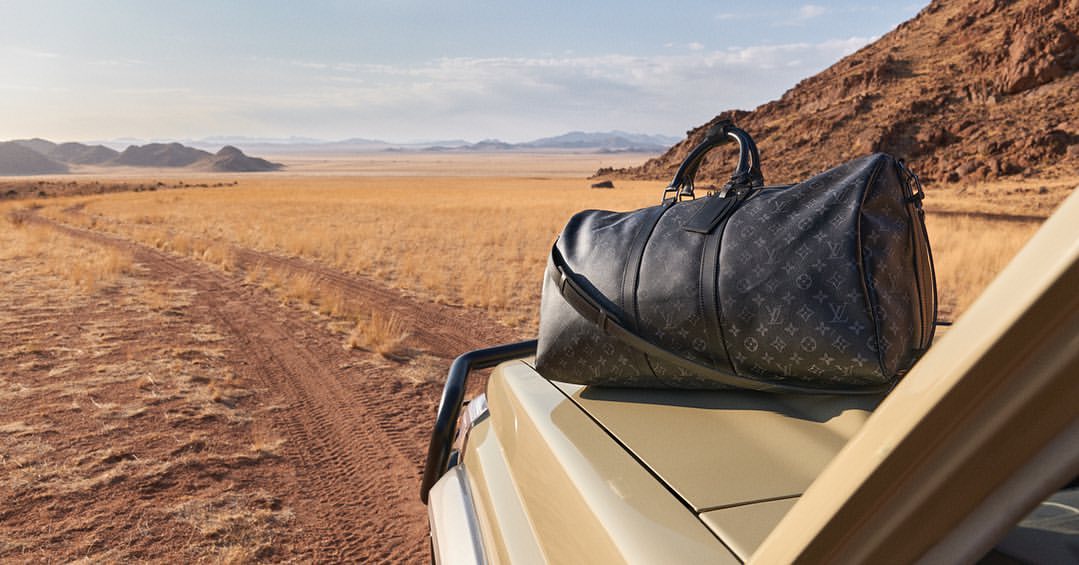 Luxury travel duffel bag by Louis Vuitton sits atop vehicle in desert landscape with mountains in the background on a clear, sunny day.