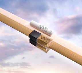 NARS foundation bottle and skincare product resting on an angled wooden beam against a sunset sky
