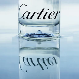 Close-up of a Cartier perfume bottle base resting on a reflective surface with soft blue lighting by Ulysse Frechelin.