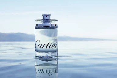 A cylindrical glass perfume bottle standing in calm shallow water with distant hazy hills under a clear sky
