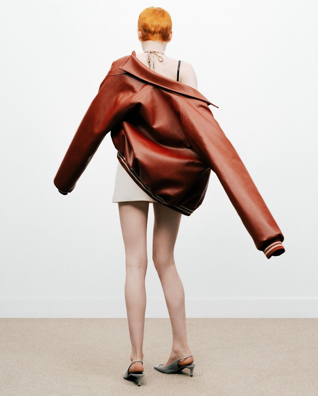 Back view of a redhead woman in a short white skirt, leather jacket on her shoulders, and gray heels, standing on a carpet against a white wall.