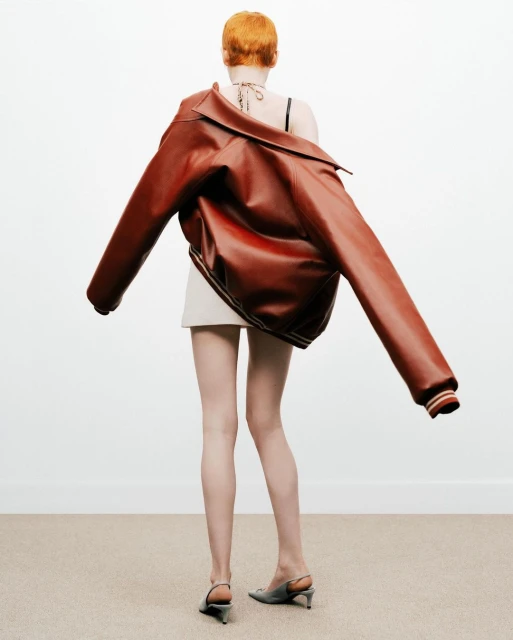 Back view of a redhead woman in a short white skirt, leather jacket on her shoulders, and gray heels, standing on a carpet against a white wall.