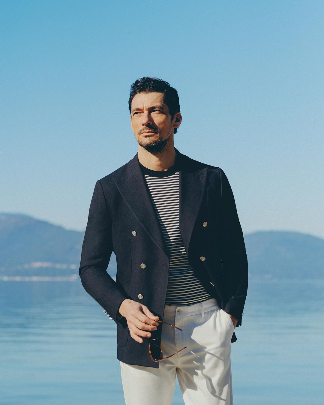 A man in a navy double-breasted blazer and white trousers standing by the water under a clear blue sky