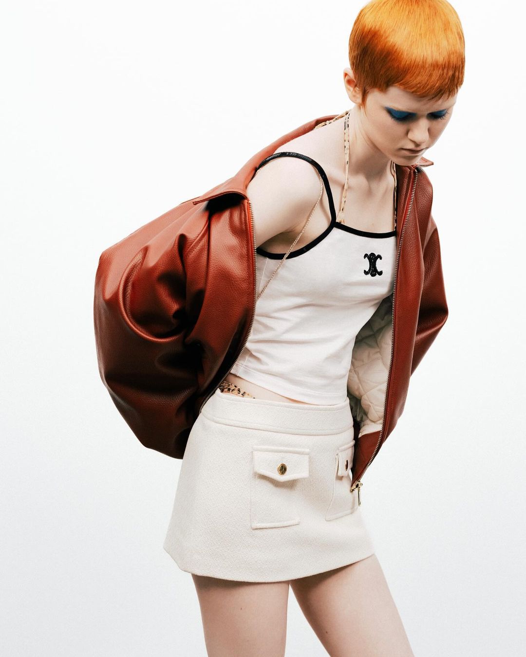 A fair-skinned woman with an orange pixie cut poses in a white tank top, cream skirt, and rust-colored leather jacket against a white backdrop.