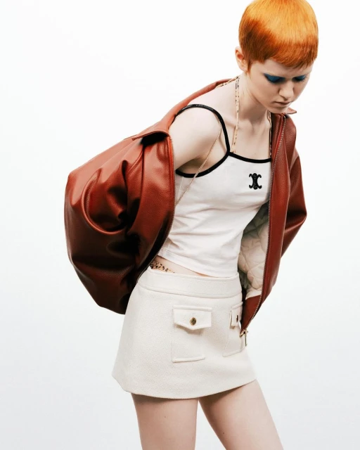 A fair-skinned woman with an orange pixie cut poses in a white tank top, cream skirt, and rust-colored leather jacket against a white backdrop.