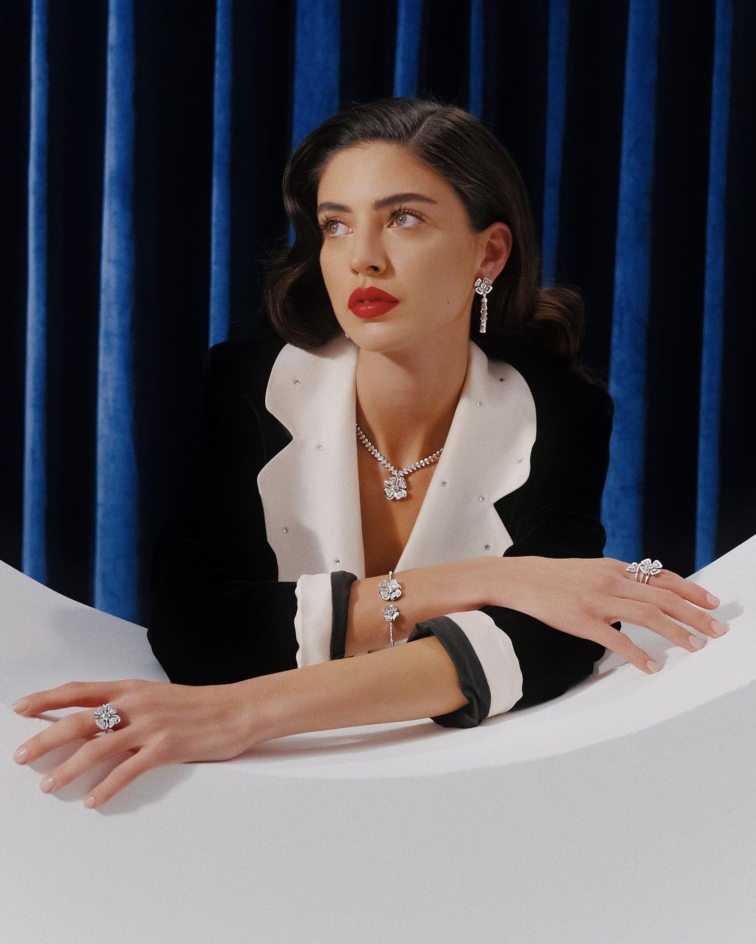 Elegant woman wearing a black and white blazer, posing with diamond jewelry against a dark blue curtain backdrop in a studio setting.