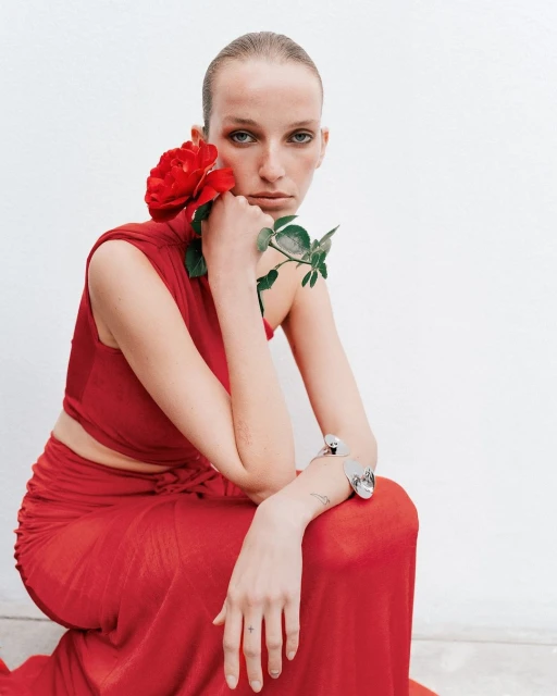 Woman in red dress poses with red rose against a white background in a fashion-style portrait with a neutral expression.