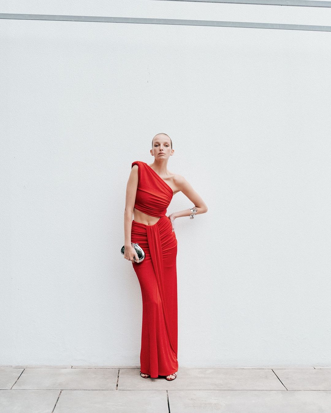 A woman poses against a white wall, wearing a red one-shoulder dress with an open midriff and carrying a silver clutch purse.