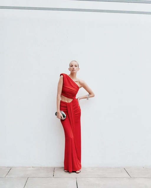 A woman poses against a white wall, wearing a red one-shoulder dress with an open midriff and carrying a silver clutch purse.