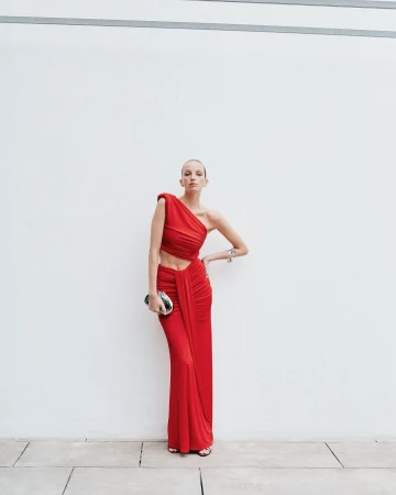 A woman poses against a white wall, wearing a red one-shoulder dress with an open midriff and carrying a silver clutch purse.