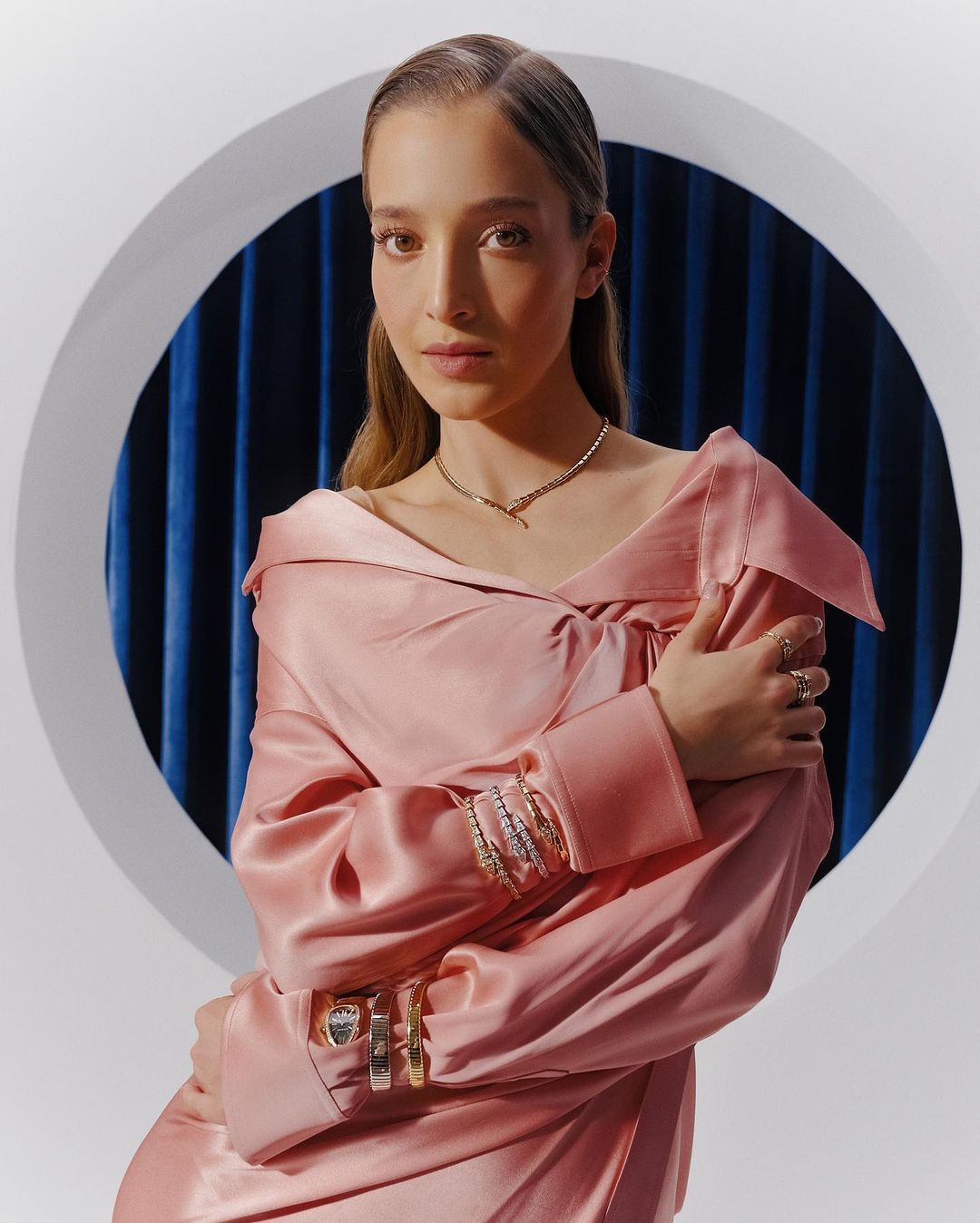 A young adult female model poses in a pink satin dress with jewelry, captured in a medium shot against a blue and white studio backdrop.