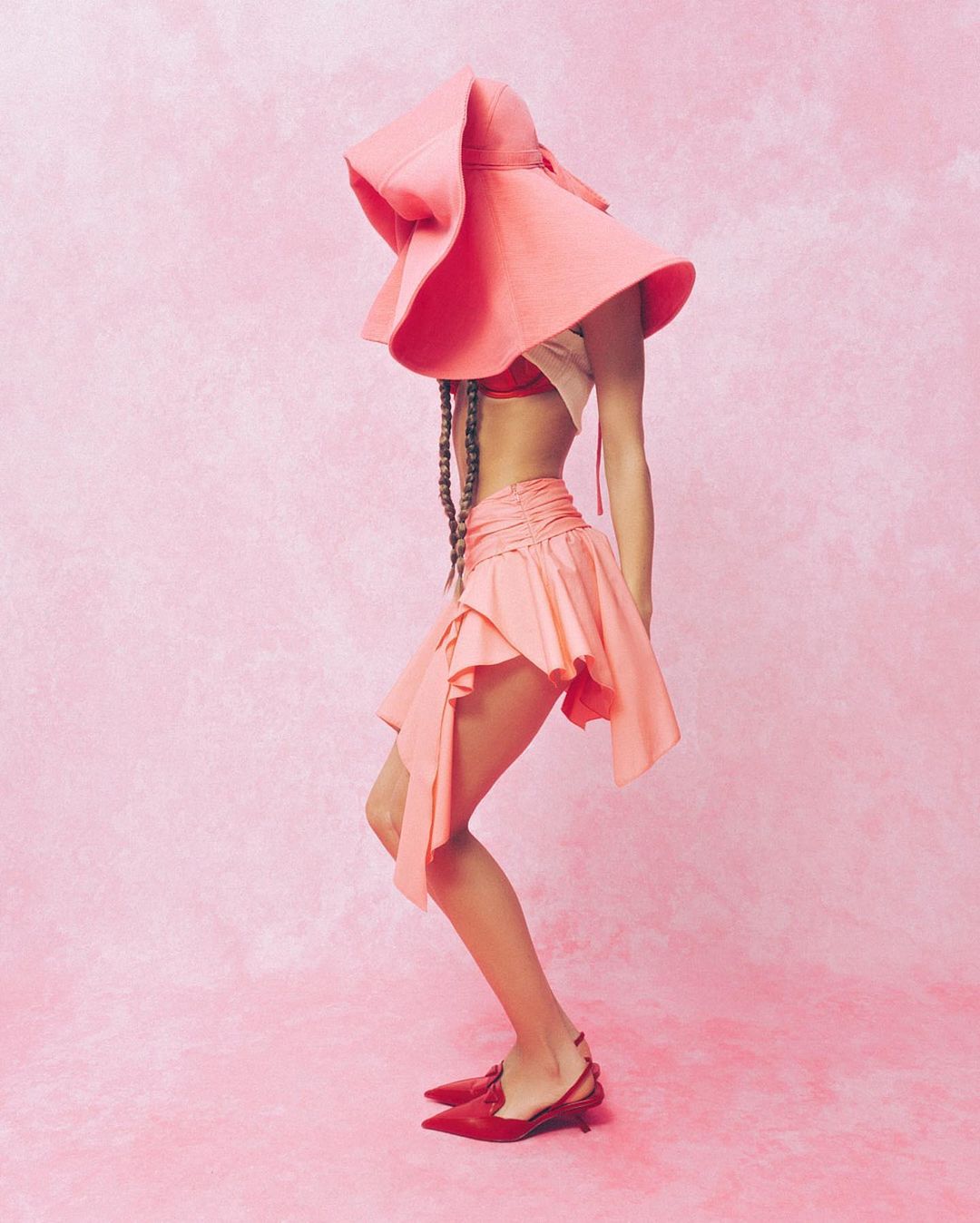 Fashionable person wearing a large pink hat, matching skirt, and red shoes, posing against a light pink textured background in studio.