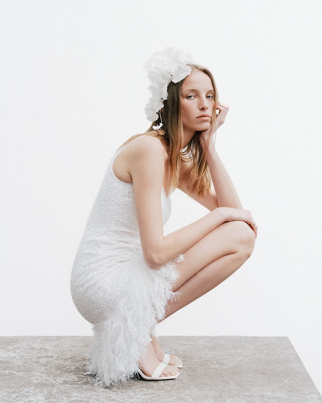 A fair-skinned woman in a white sequin dress poses in a crouched position with a white floral headpiece against a white background.