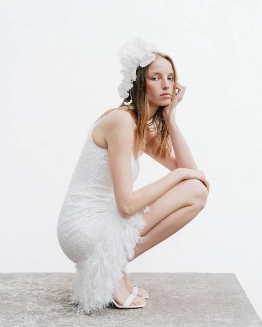 A fair-skinned woman in a white sequin dress poses in a crouched position with a white floral headpiece against a white background.