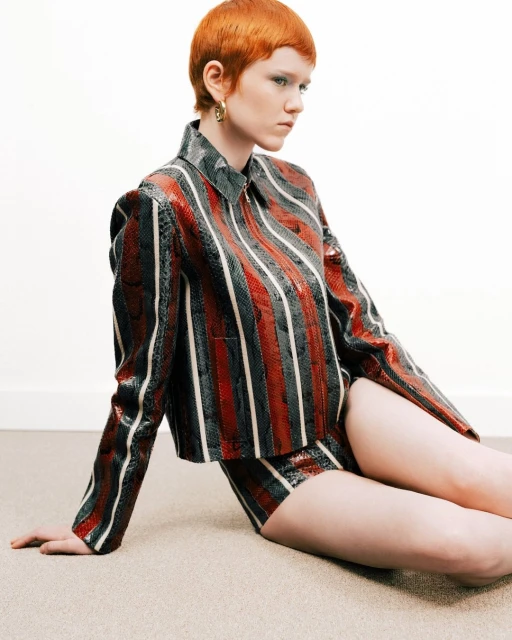 A red-haired woman sits on the floor in a striped jacket and shorts. Soft daylight illuminates the indoor fashion portrait.