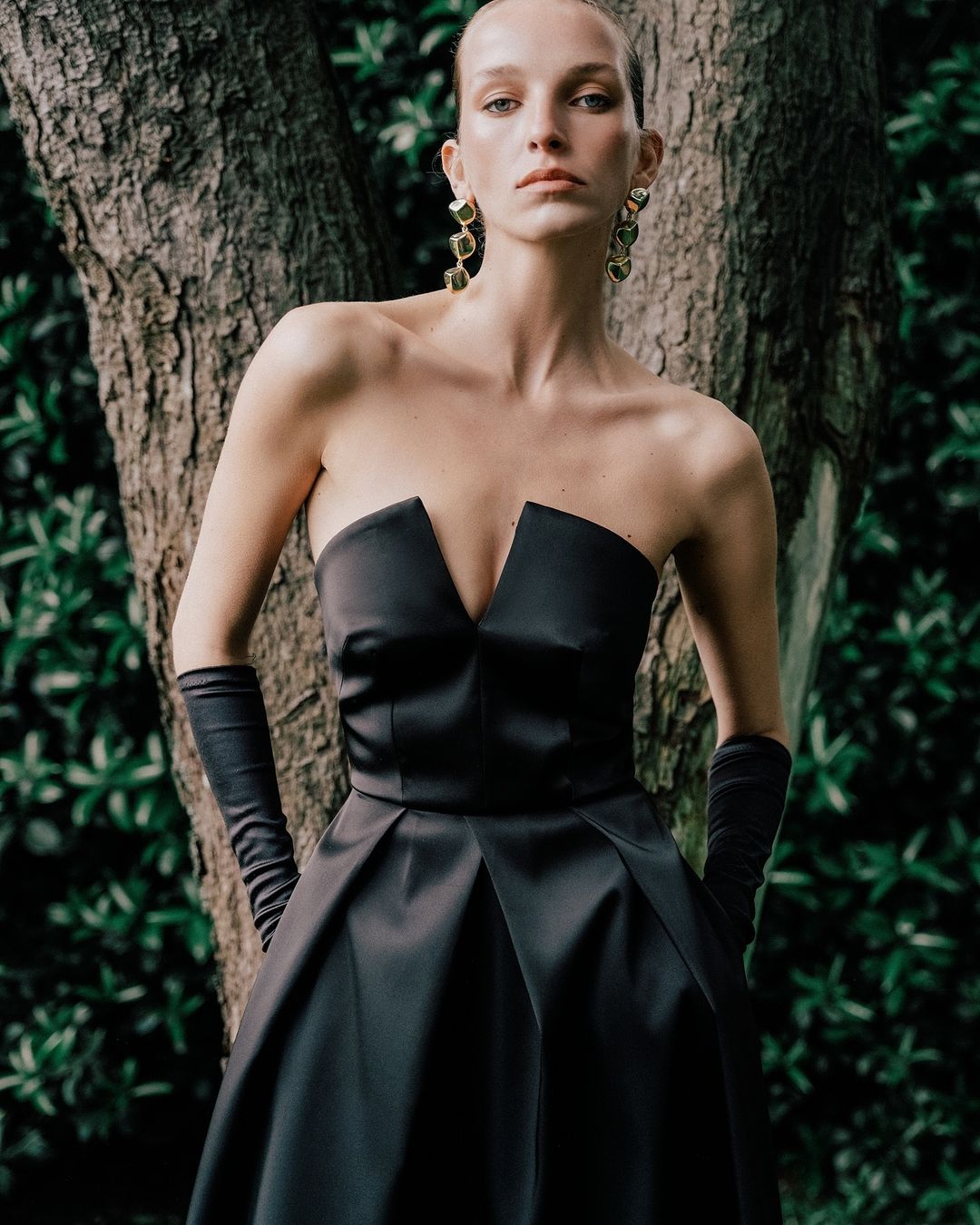 Elegant woman in strapless black dress posing near tree. Fashion shoot with model in satin gown and velvet gloves. Stylish outdoor portrait.