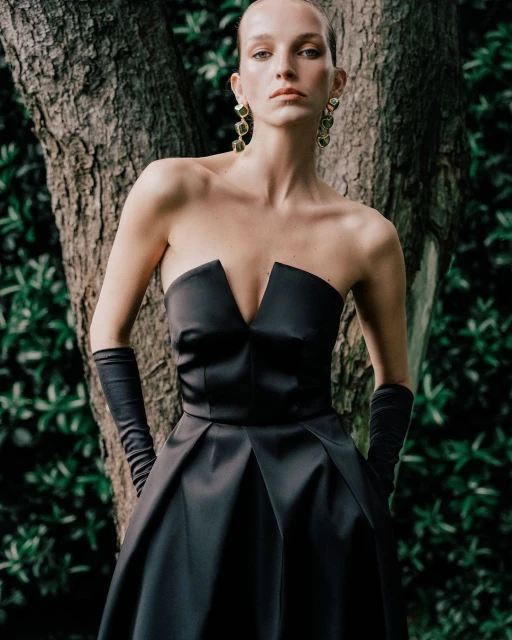 Elegant woman in strapless black dress posing near tree. Fashion shoot with model in satin gown and velvet gloves. Stylish outdoor portrait.