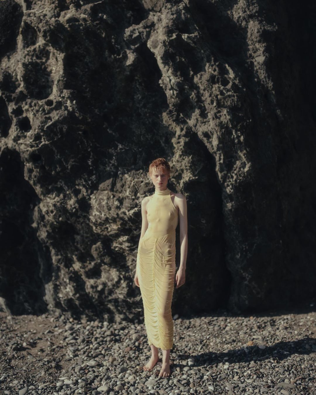 A red-haired model in a yellow dress stands barefoot on a pebbled beach in front of a large rock formation.