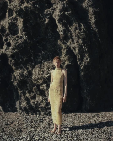 A red-haired model in a yellow dress stands barefoot on a pebbled beach in front of a large rock formation.