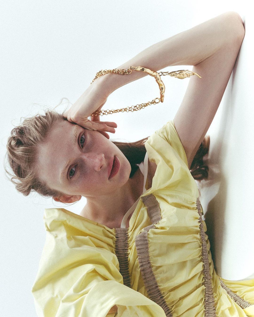 Woman in yellow dress poses with gold necklace in a studio, looking at the camera. Fashion editorial, portrait. High-key lighting.