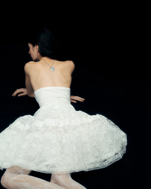 Woman in white dress and lace tights poses against a black backdrop, showcasing fashion and elegance, with a shallow depth of field.