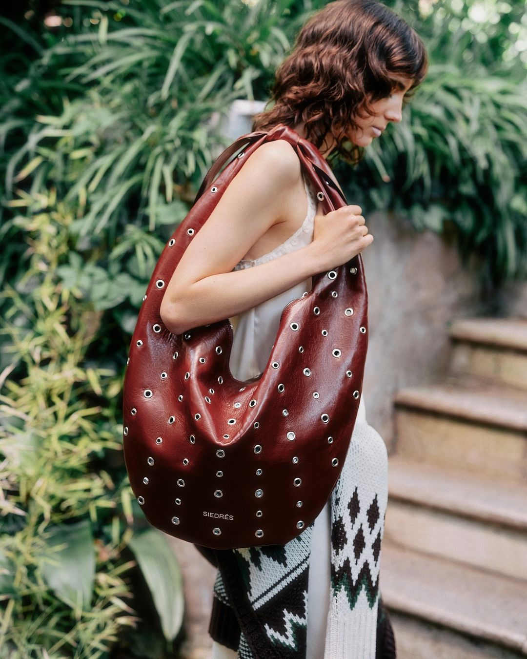 A woman carries a large, burgundy leather tote bag with silver grommets, walking on stone steps surrounded by lush green foliage.