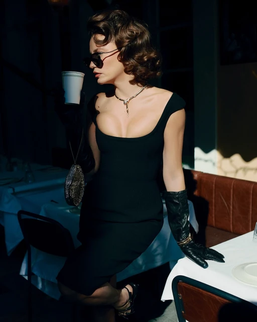 Elegant woman in a black dress, opera gloves, and sunglasses poses indoors with a purse and coffee cup, bathed in moody, cinematic light.