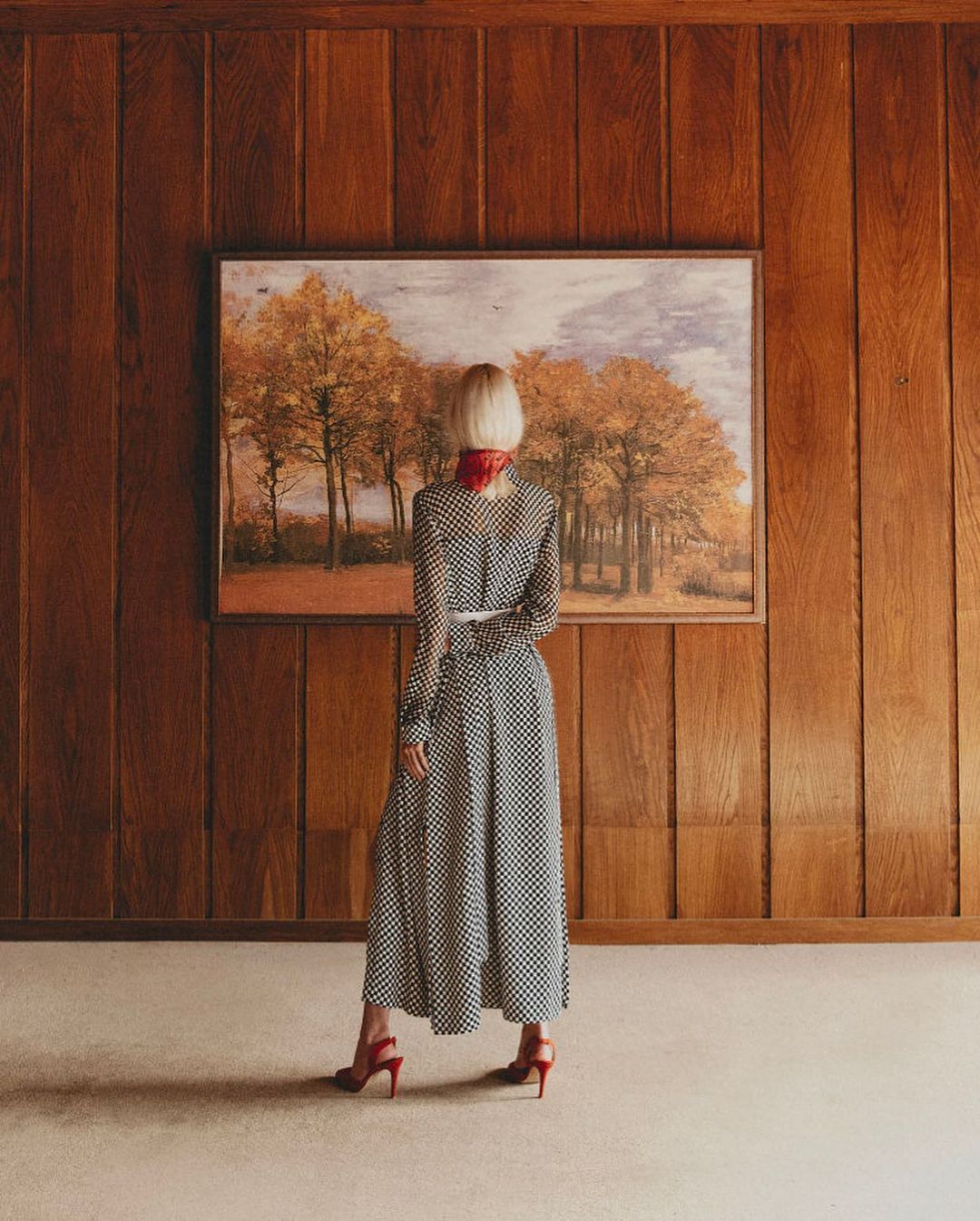 A woman with short blonde hair wearing a long polka dot dress and red heels stands in front of a landscape painting in a wood-paneled room.