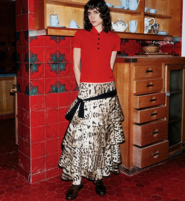Young woman poses indoors in a red short-sleeve shirt and tiered animal-print skirt, standing in a kitchen with red tile walls.