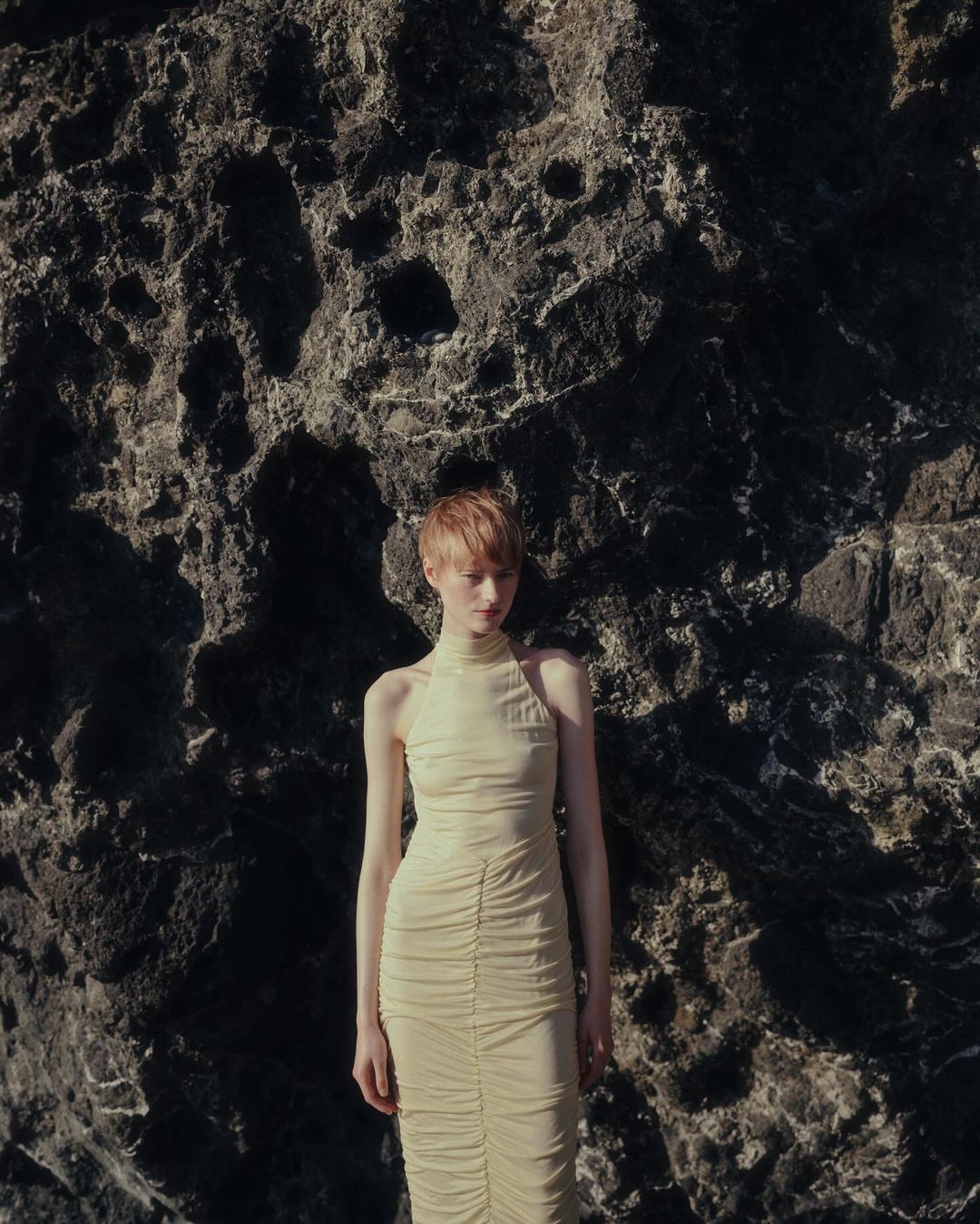 Woman in a long beige dress posing against a textured grey rock formation in natural light with a shallow depth of field.