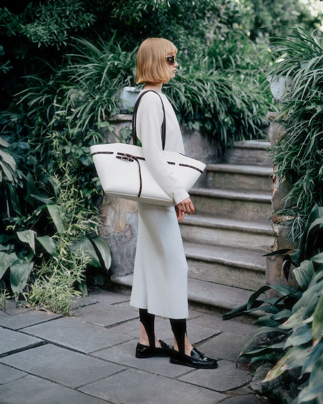 Woman with short blonde hair wearing a white long-sleeve dress and holding a large white tote bag standing outdoors near stone steps.