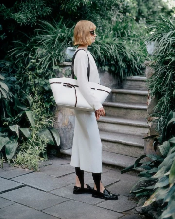 Woman with short blonde hair wearing a white long-sleeve dress and holding a large white tote bag standing outdoors near stone steps.