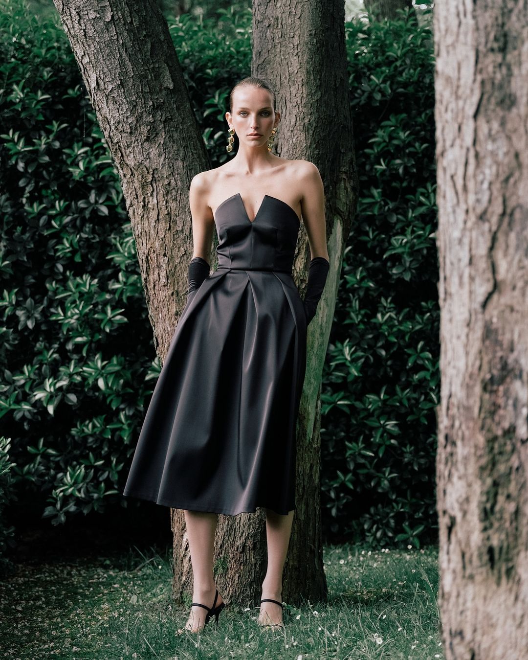 Elegant woman in a black strapless dress stands next to trees in a green outdoor setting, showcasing a classic and sophisticated fashion style.