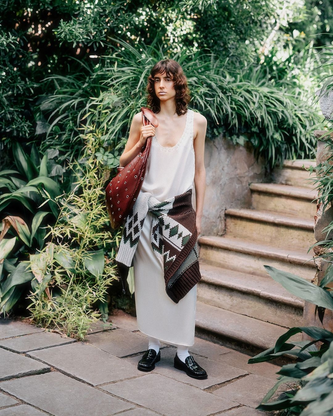 A woman in a slip dress and flats stands on a stone path lined with lush greenery, carrying a large leather bag and tied sweater.