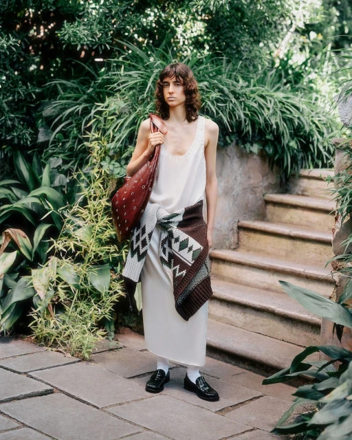A woman in a slip dress and flats stands on a stone path lined with lush greenery, carrying a large leather bag and tied sweater.