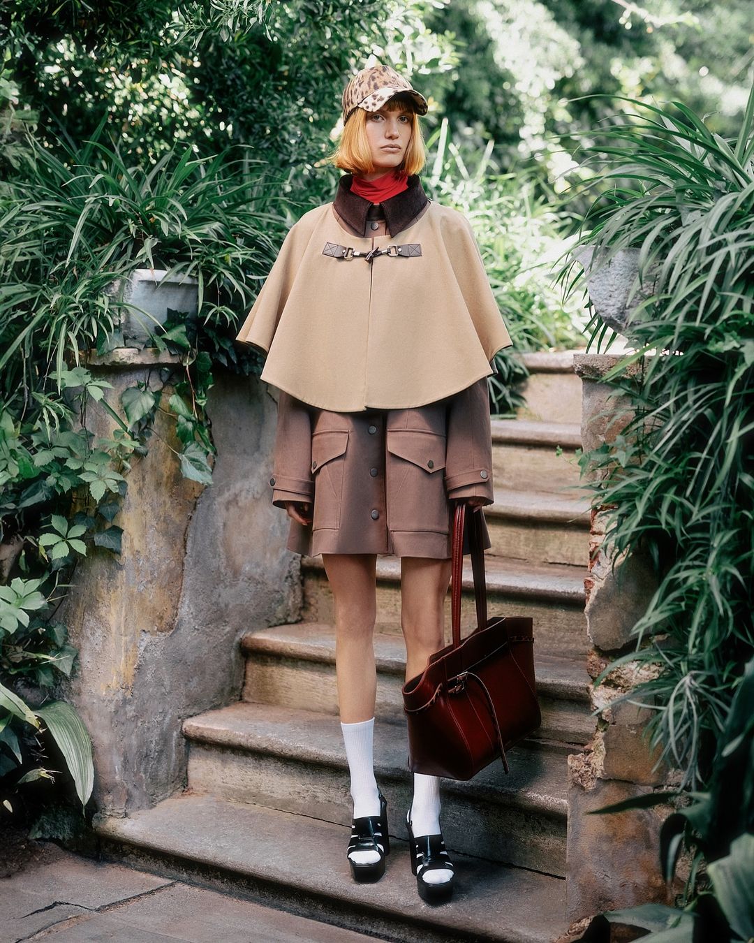 Fashionable woman in a tan cape and matching hat poses on stone steps surrounded by greenery, carrying a dark brown leather tote bag.