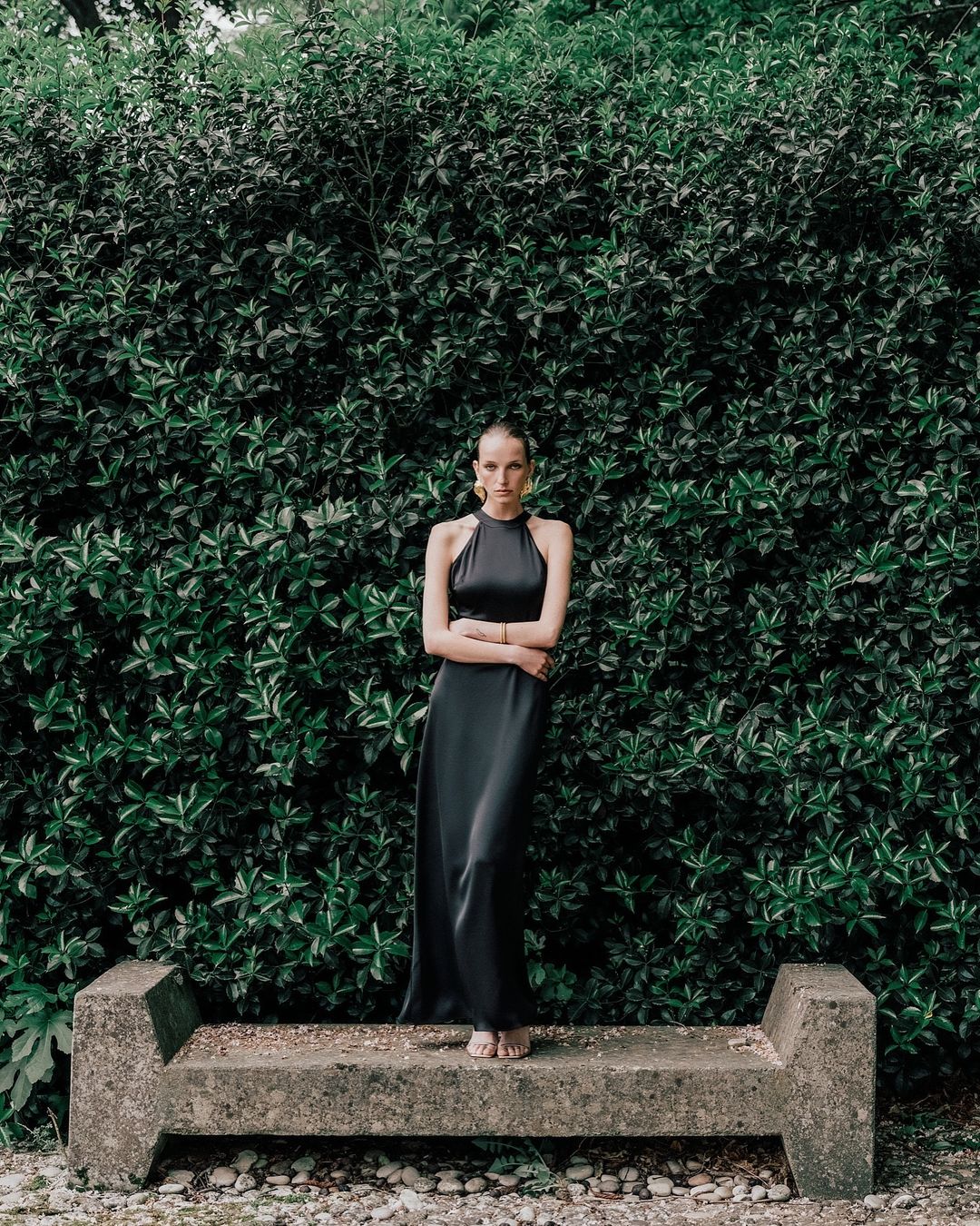 A barefoot woman in a long black halter dress stands on a concrete bench with arms crossed in front of a large green leafy wall.