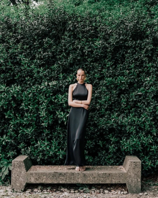 A barefoot woman in a long black halter dress stands on a concrete bench with arms crossed in front of a large green leafy wall.