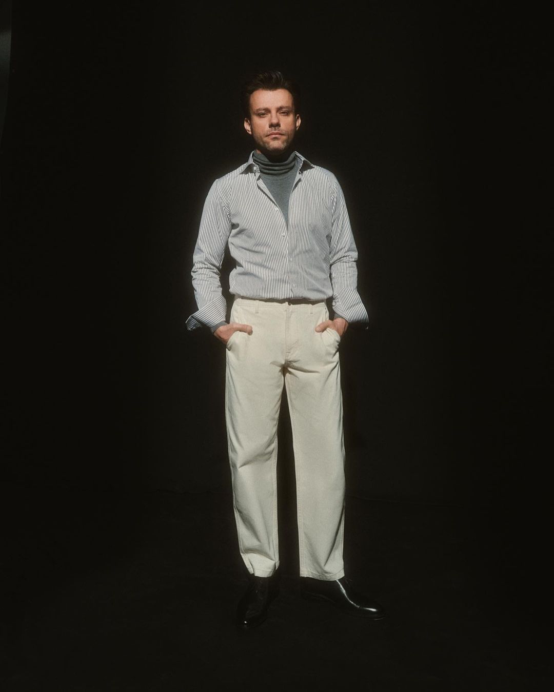 Full-length portrait of a stylish adult male in a striped shirt and off-white trousers, set against a dark backdrop in a studio setting.