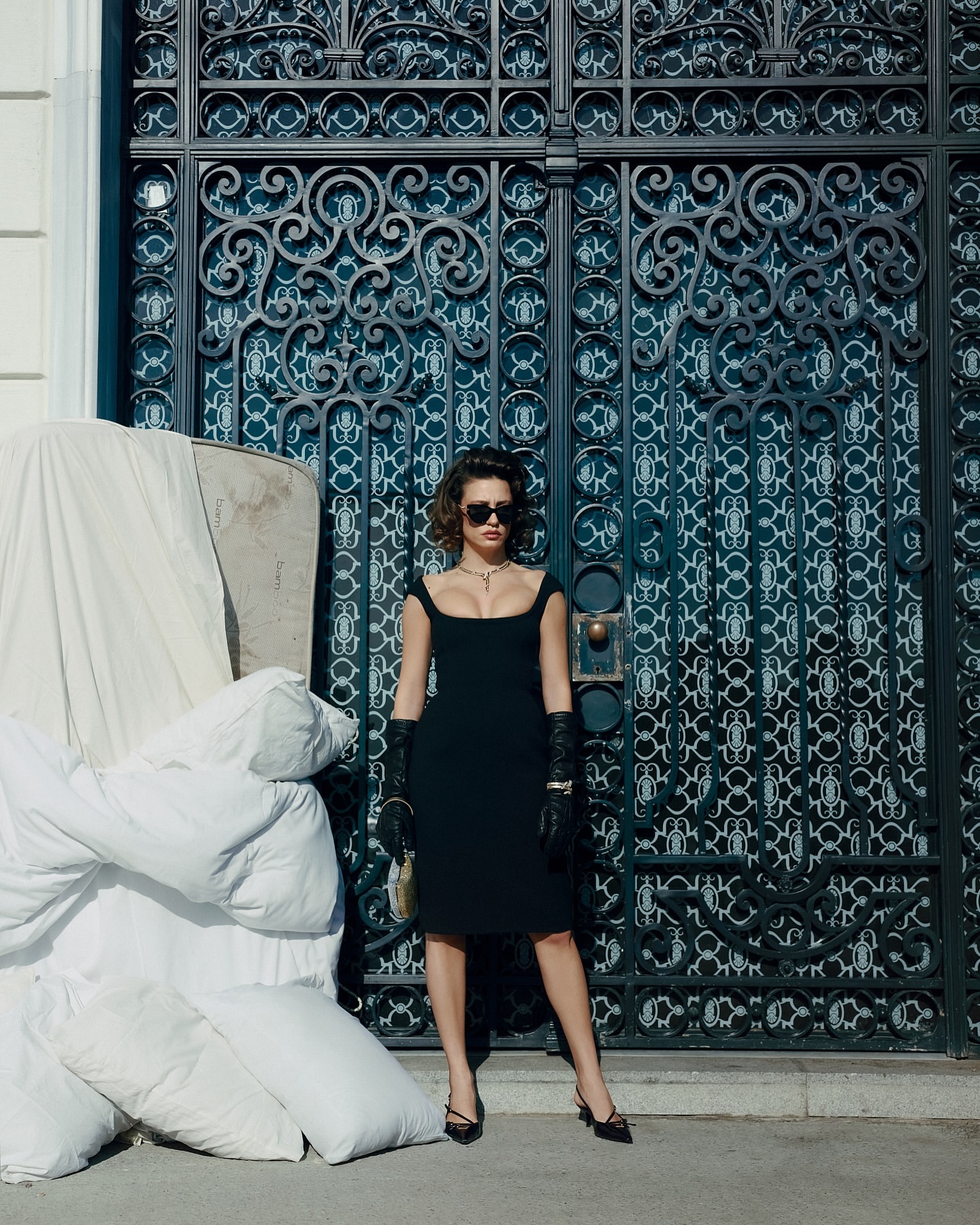 A woman poses in a black dress, gloves, and heels, against an ornamental black gate with a pile of white fabric or bedding beside her.